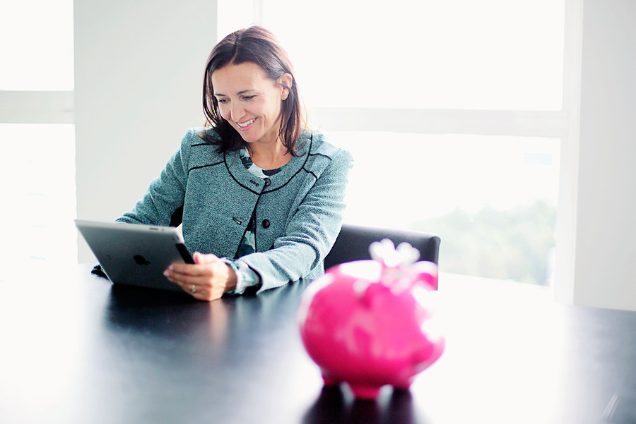 Een vrouw kijkt op haar tablet en op tafel staat een roze spaarvarken